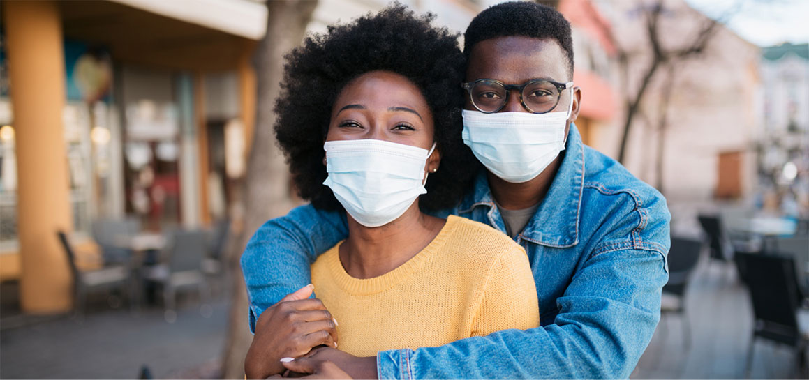 Wearing their masks, an African American husband and his wife embrace.