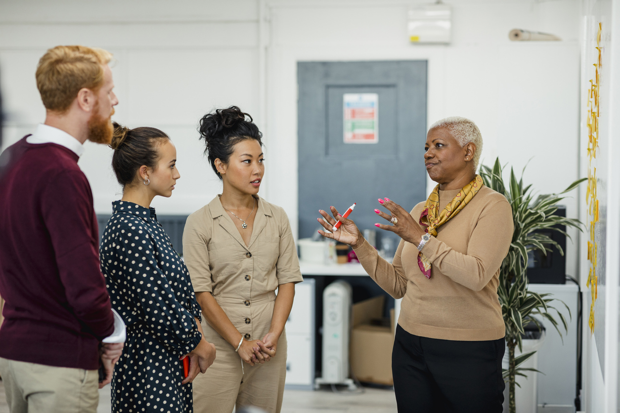 Case Management Strategies & Coaching – A woman stands at a whiteboard holding a marker while talking to three other people.