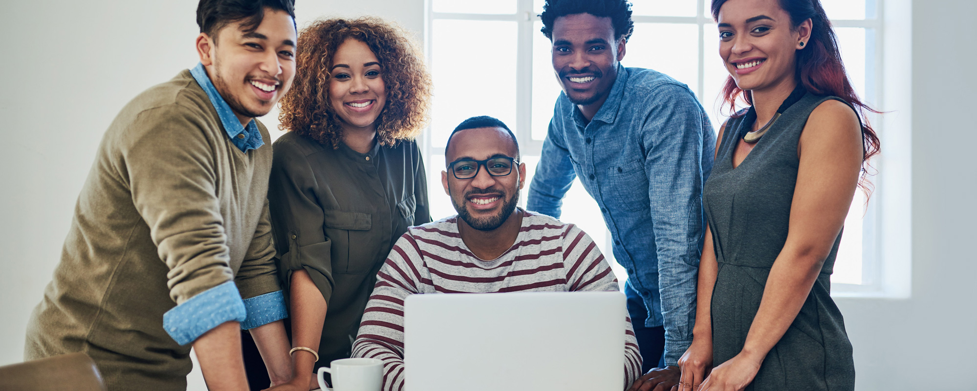 A group of five employees smiling, one employee is sitting down and others standing.